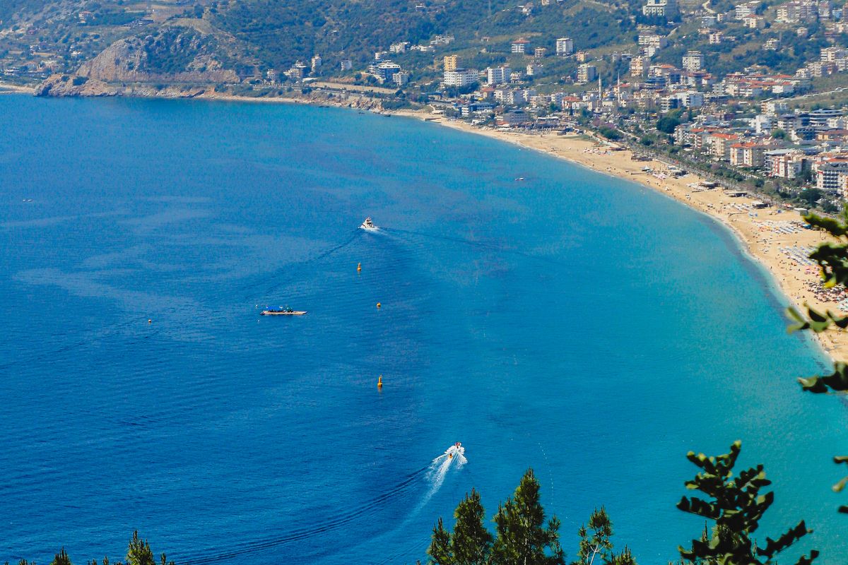The long curve of Cleopatra Beach in Alanya seen from the castle slope, with turquoise water, a headland and the town behind
