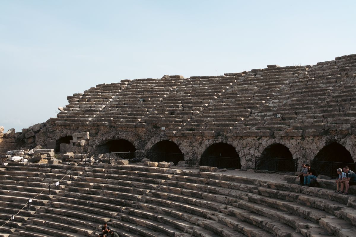 The tiered stone seating and arched vaults of the Roman theatre at Side, with a few visitors sitting on the steps